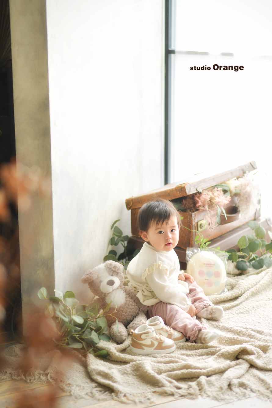 1歳女の子が一升餅とファーストトイのくまさんのぬいぐるみとファーストシューズと共に写っている様子